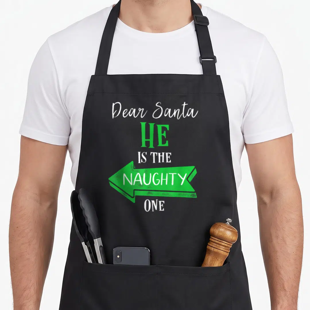 Couple chef outfit apron - Stacked text Dear Santa HE IS THE NAUGHTY ONE with green arrow pointing left — shown on a black cotton canvas apron against a white background