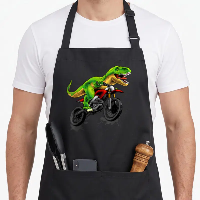 Dirt dinosaur apron - Green T-Rex riding red dirt bike jumping cartoon art — shown on a black cotton canvas apron against a white background