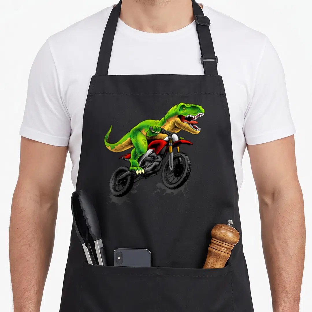 Dirt dinosaur apron - Green T-Rex riding red dirt bike jumping cartoon art — shown on a black cotton canvas apron against a white background