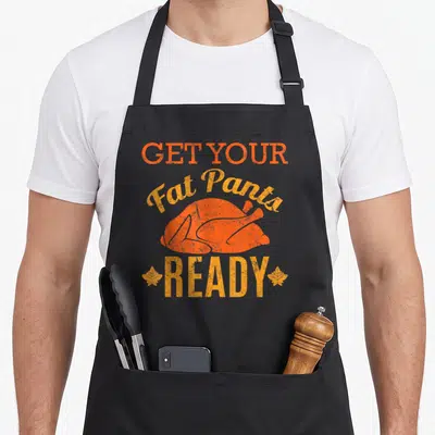 Pant fat man in apron - Distressed orange text Get Your Fat Pants Ready over roasted turkey with two leaves — shown on a black cotton canvas apron against a white background