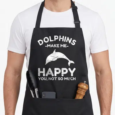 Funny dolphin dad apron - White distressed dolphin graphic with text about making the wearer happy but not others — shown on a black cotton canvas apron against a white background