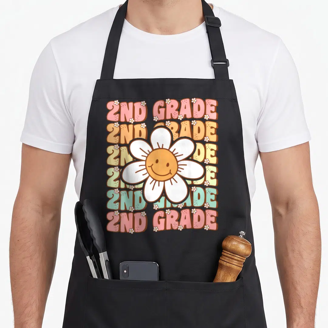 Groovy 2nd apron - Six rows of colorful 2ND GRADE text with a central smiling white daisy and small scattered flowers — shown on a black cotton canvas apron against a white background
