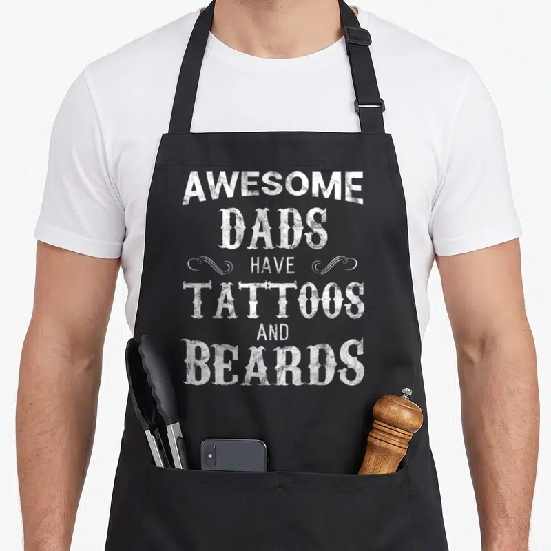 Father tattoo aprons - White stacked text reading AWESOME DADS HAVE TATTOOS AND BEARDS with decorative swirls — shown on a black cotton canvas apron against a white background