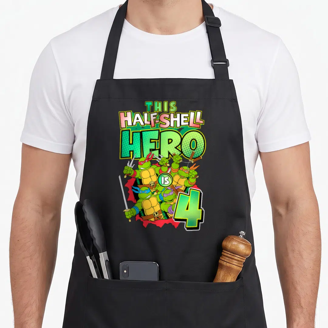 Hero teenage mutant ninja apron - Four turtles bursting red background text This Half-Shell Hero Is 4 cartoon style — shown on a black cotton canvas apron against a white background