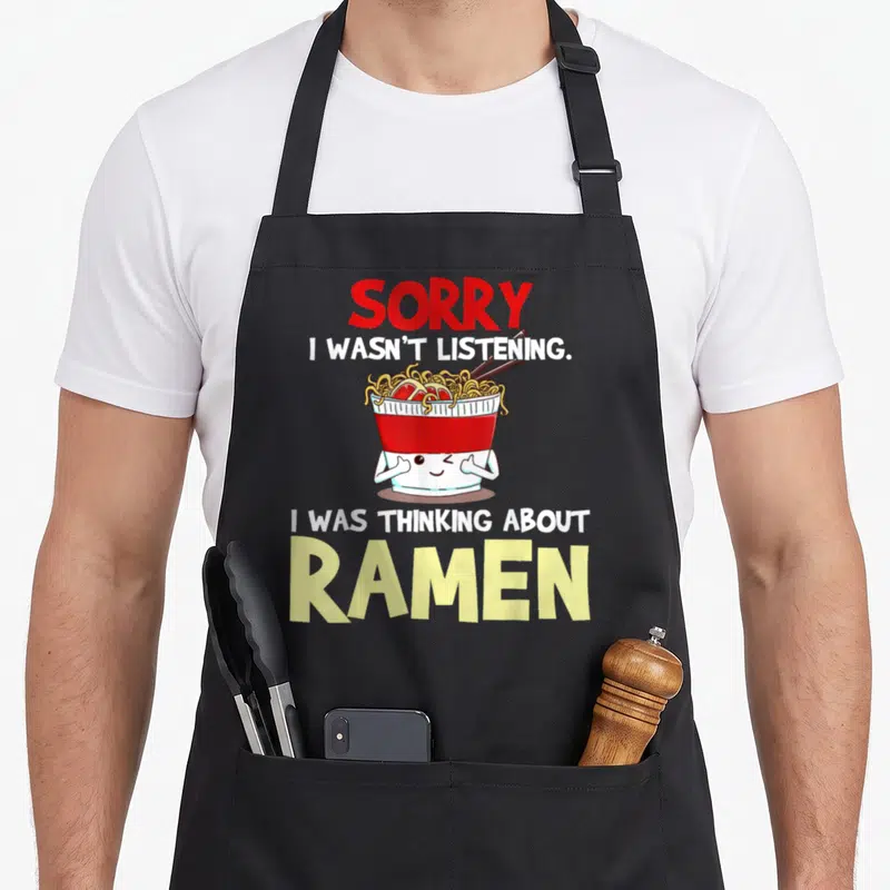 Japanese ramen noodles apron - Cartoon ramen bowl with arms winking between red and yellow text reading sorry i wasn t — shown on a black cotton canvas apron against a white background