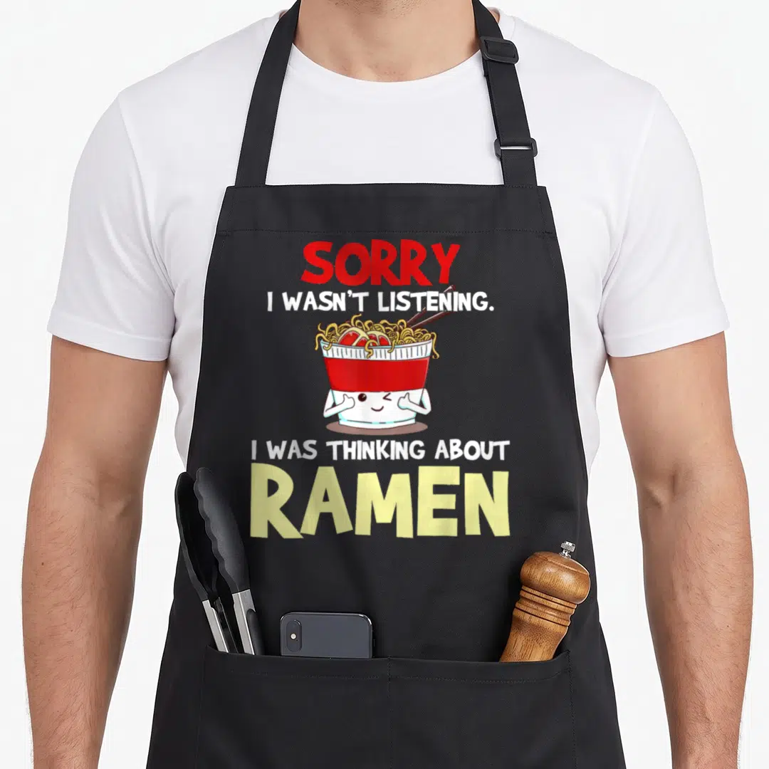 Japanese ramen noodles apron - Cartoon ramen bowl with arms winking between red and yellow text reading sorry i wasn t — shown on a black cotton canvas apron against a white background