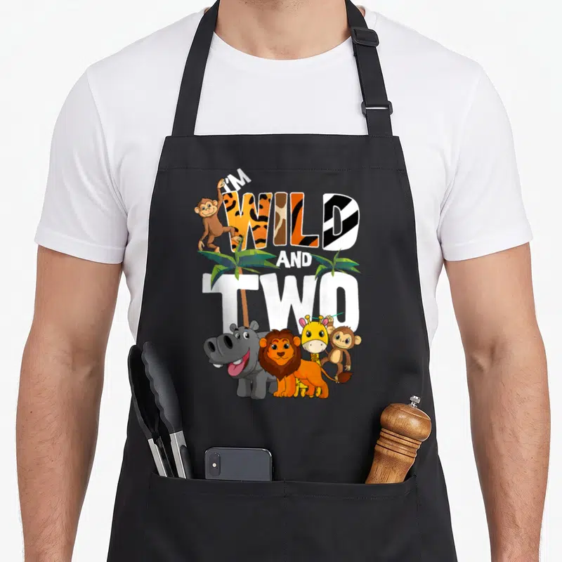 Bday 2nd apron - I'M WILD AND TWO text with cartoon monkeys, lion, hippo, giraffe, and palm trees in orange and green — shown on a black cotton canvas apron against a white background