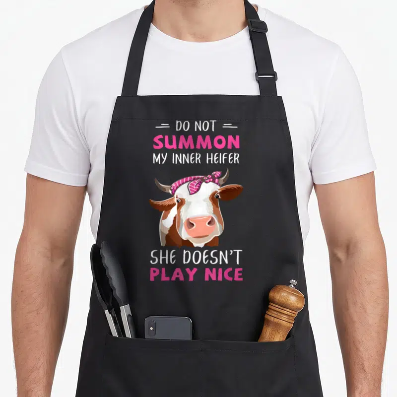 Summon cow print aprons - Cartoon cow with pink bandana and text Do Not Summon My Inner Heifer She Doesn't Play Nice — shown on a black cotton canvas apron against a white background