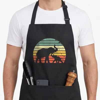Sunset elephant print apron - Grey adult and baby elephant silhouettes against a striped retro sun in green, yellow — shown on a black cotton canvas apron against a white background