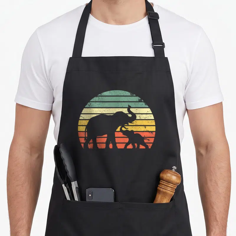 Sunset elephant print apron - Grey adult and baby elephant silhouettes against a striped retro sun in green, yellow — shown on a black cotton canvas apron against a white background