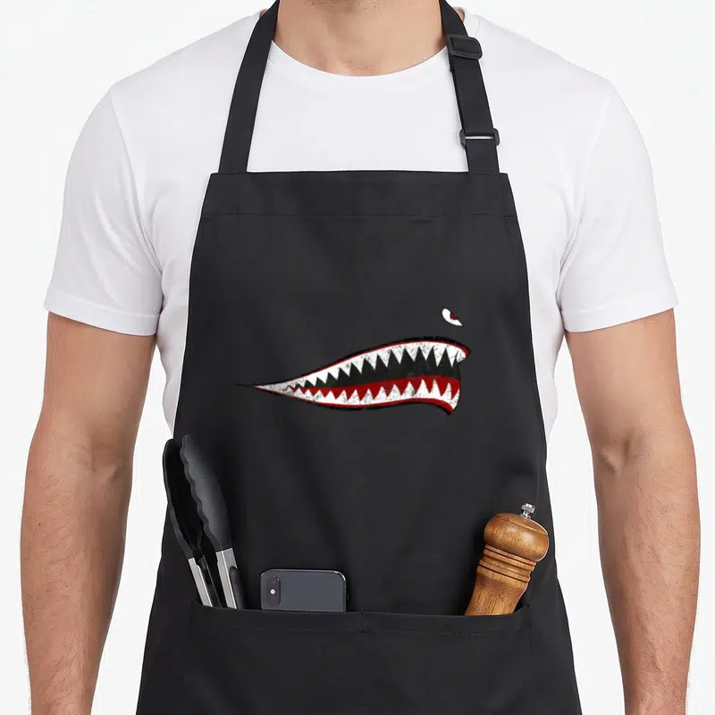 Shark ww2 apron - Distressed shark mouth with sharp white teeth, red accents, and a single floating eye looking down — shown on a black cotton canvas apron against a white background