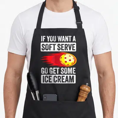 Pickleball ice to serve man apron - Yellow ball with red trail and text about soft serve ice cream — shown on a black cotton canvas apron against a white background