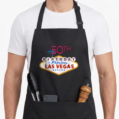 50th las birthday apron - Retro neon sign graphic with 50TH BIRTHDAY IN Fabulous LAS VEGAS NEVADA text and yellow bulbs — shown on a black cotton canvas apron against a white background