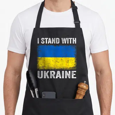 Support ukrainian aprons - Distressed blue and yellow flag flanked by white text I STAND WITH UKRAINE on black fabric — shown on a black cotton canvas apron against a white background