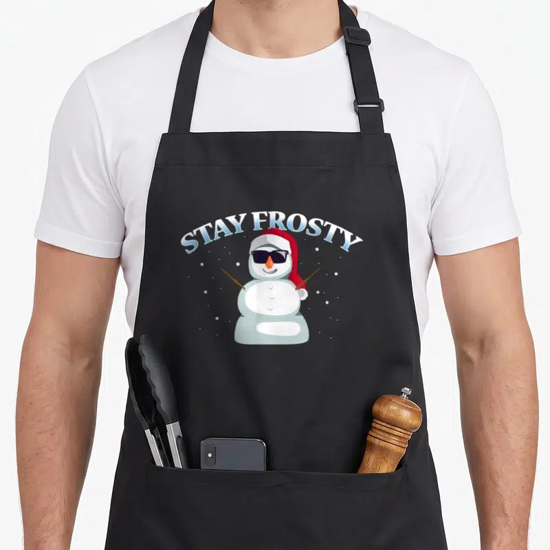 Stay merry christmas apron - Cartoon snowman with sunglasses and Santa hat under arched blue text STAY FROSTY — shown on a black cotton canvas apron against a white background