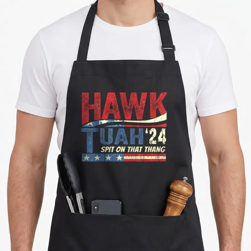 Spit hawk tuah apron - Red white blue distressed text HAWK TUAH '24 with flag wave and strip elements — shown on a black cotton canvas apron against a white background
