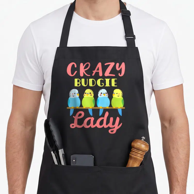 Parakeet pet bird apron - Four colorful budgies perched on brown branch below CRAZY BUDGIE Lady text — shown on a black cotton canvas apron against a white background