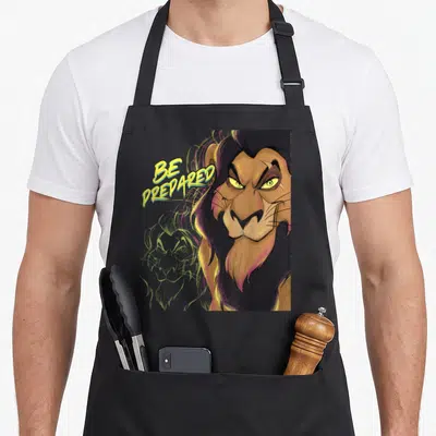 Scar halloween apron - Brown lion head with yellow BE PREDARED text and faint green sketch background — shown on a black cotton canvas apron against a white background
