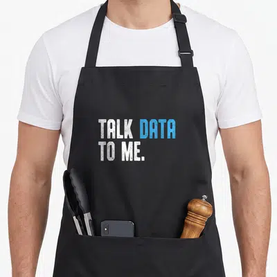 Talk data apron - TALK DATA TO ME text in white and blue stacked typography — shown on a black cotton canvas apron against a white background