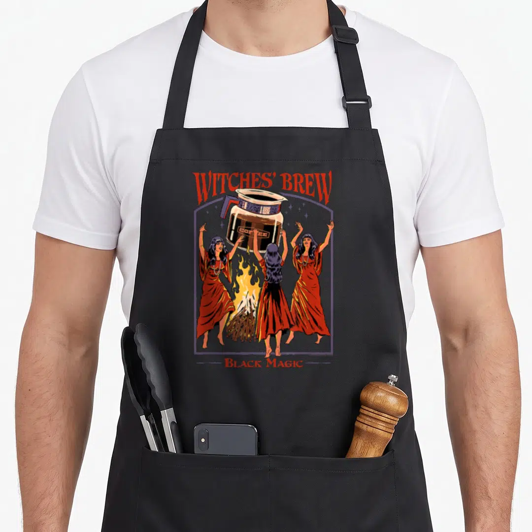 Rhode magic apron - Three cartoon witches in red dresses levitating a coffee pot over a fire under arched WITCHES' BREW — shown on a black cotton canvas apron against a white background
