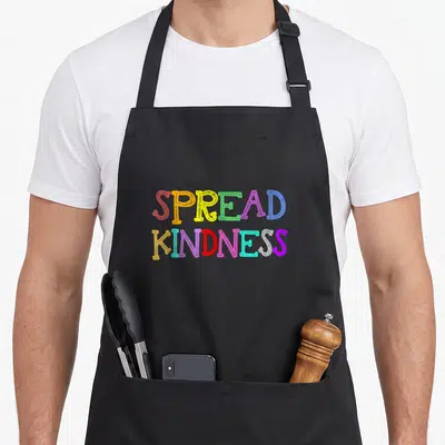 Anti bullying prevention apron - Stacked SPREAD KINDNESS text in red yellow green blue purple orange pink and cyan — shown on a black cotton canvas apron against a white background