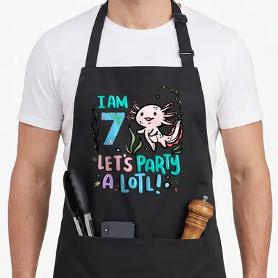7th axolotl birthday apron - Cartoon pink axolotl with I AM 7 LET'S PARTY A LOTL text and teal number 7 — shown on a black cotton canvas apron against a white background