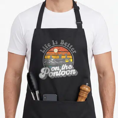 Better houseboat apron - Retro sunset circle with pontoon silhouette and Life Is Better on the Pontoon text — shown on a black cotton canvas apron against a white background