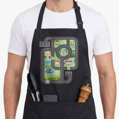 Play track apron - Cartoon town map with grey roads, green areas, and buildings labeled Gas Station, Market, and School — shown on a black cotton canvas apron against a white background