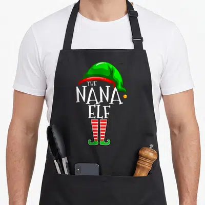 Family nana aprons - Green elf hat and red striped legs framing white The Nana Elf text in cartoon style — shown on a black cotton canvas apron against a white background