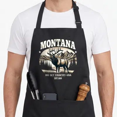 Rocky country apron - Retro elk illustration with arched Montana text and mountain landscape in earth tones — shown on a black cotton canvas apron against a white background