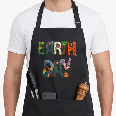 Earth april apron - EARTH DAY letters filled with flowers, birds, coral, and trees in green orange blue red purple — shown on a black cotton canvas apron against a white background