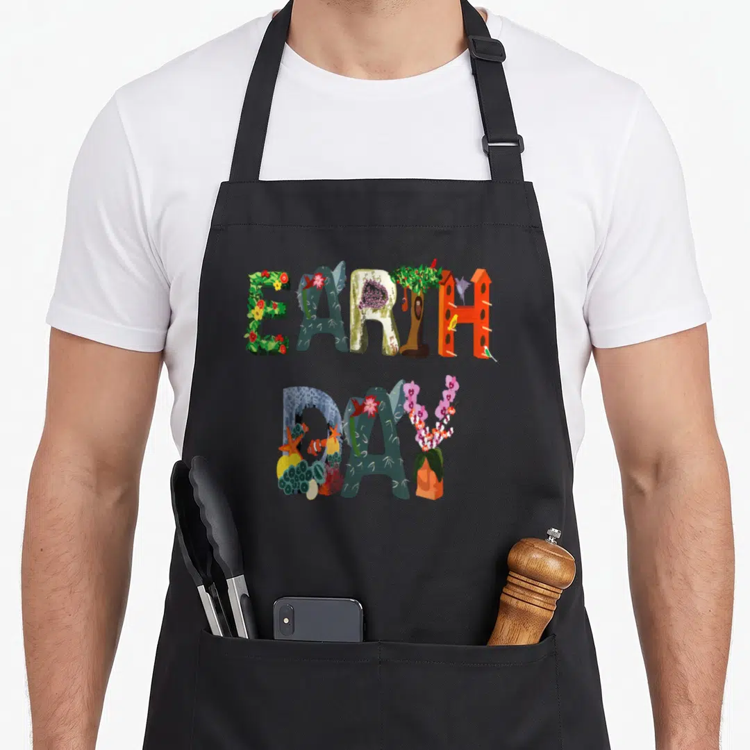 Earth april apron - EARTH DAY letters filled with flowers, birds, coral, and trees in green orange blue red purple — shown on a black cotton canvas apron against a white background