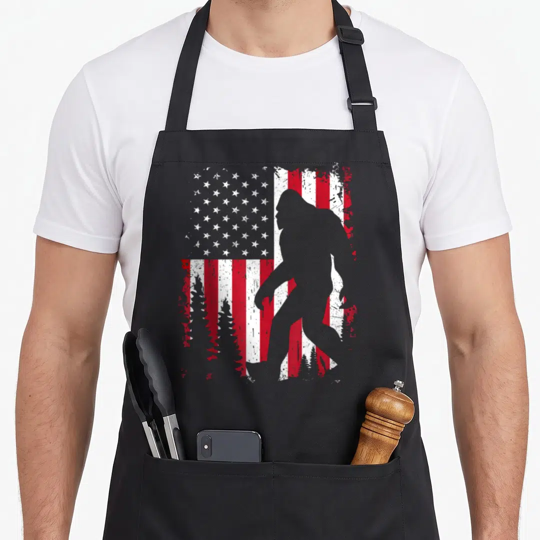 Bigfoot 4th of july apron - Grey Bigfoot silhouette over distressed red white flag with three pine trees — shown on a black cotton canvas apron against a white background