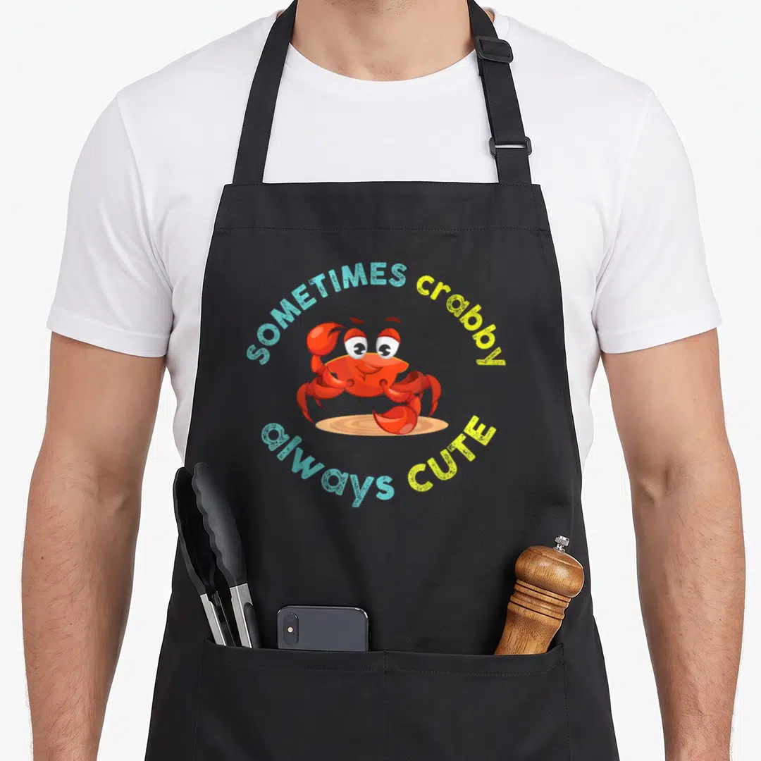 Sometime crab apron - Red cartoon crab on sand with SOMETIMES crabby always CUTE text in blue and yellow arcs — shown on a black cotton canvas apron against a white background