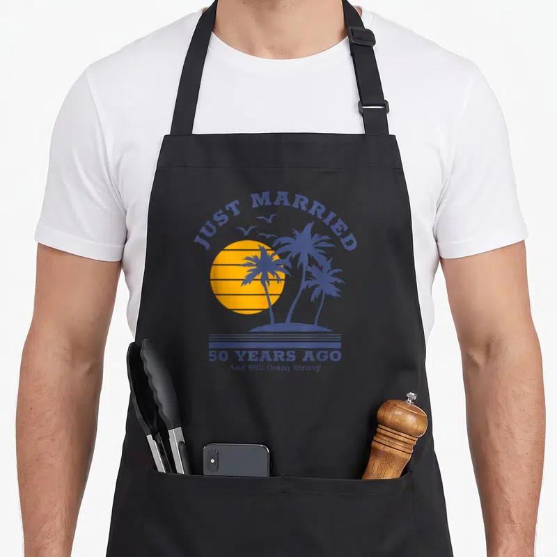 Ago wedding apron - JUST MARRIED text over yellow sun and purple palm trees with 50 YEARS AGO caption — shown on a black cotton canvas apron against a white background