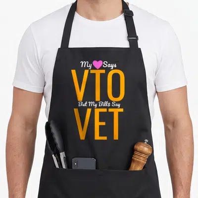 Vto employee aprons - Orange VTO and VET text with pink heart and white script saying My Says But My Bill$ Say — shown on a black cotton canvas apron against a white background