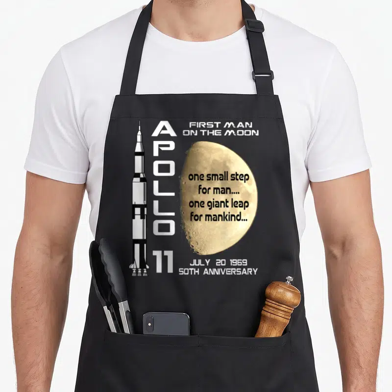 July apollo apron - Apollo 11 rocket and moon typography design with 1969 anniversary text — shown on a black cotton canvas apron against a white background