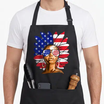 Afro 4th of july apron - Distressed portrait with feathered headdress and flag sunglasses in red white blue brown gold — shown on a black cotton canvas apron against a white background