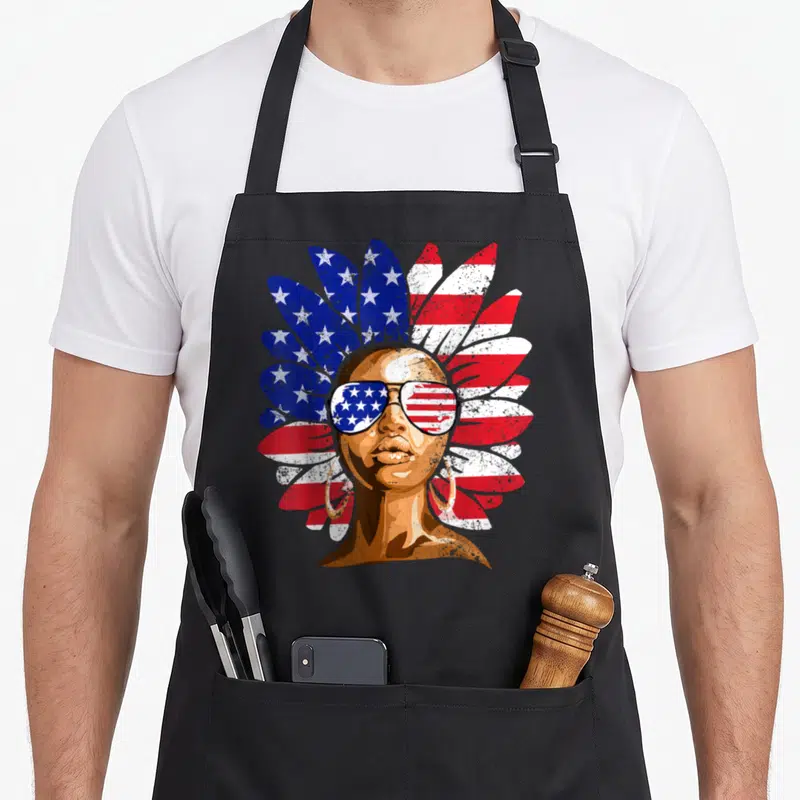 Afro 4th of july apron - Distressed portrait with feathered headdress and flag sunglasses in red white blue brown gold — shown on a black cotton canvas apron against a white background