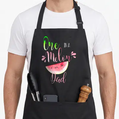 Melon birthday apron - Watercolor watermelon slice with 'One IN A Melon Dad' text in pink and green script — shown on a black cotton canvas apron against a white background