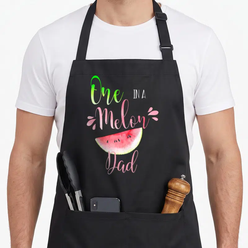 Melon birthday apron - Watercolor watermelon slice with 'One IN A Melon Dad' text in pink and green script — shown on a black cotton canvas apron against a white background