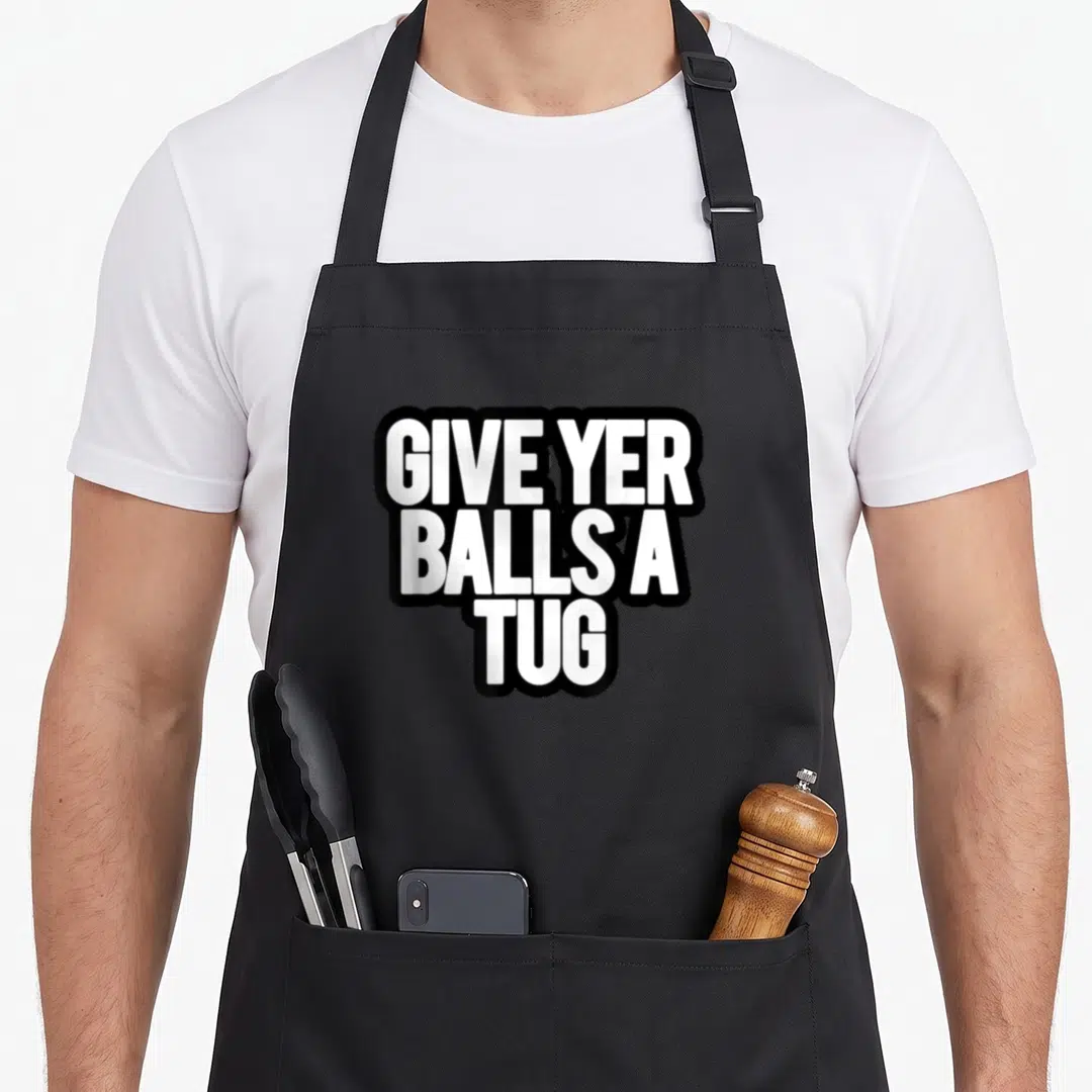 Give ostriches ostrich apron - Stacked white text GIVE YER BALLS A TUG with thick black outline — shown on a black cotton canvas apron against a white background