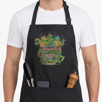 Pizza apron for mom - Four turtles around manhole cover with TEENAGE MUTANT NINJA TURTLES text and pizza — shown on a black cotton canvas apron against a white background
