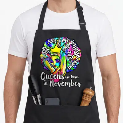 Saggittarius queen apron - Colorful cartoon Afro woman with crown surrounded by affirmations and 'Queens are born — shown on a black cotton canvas apron against a white background
