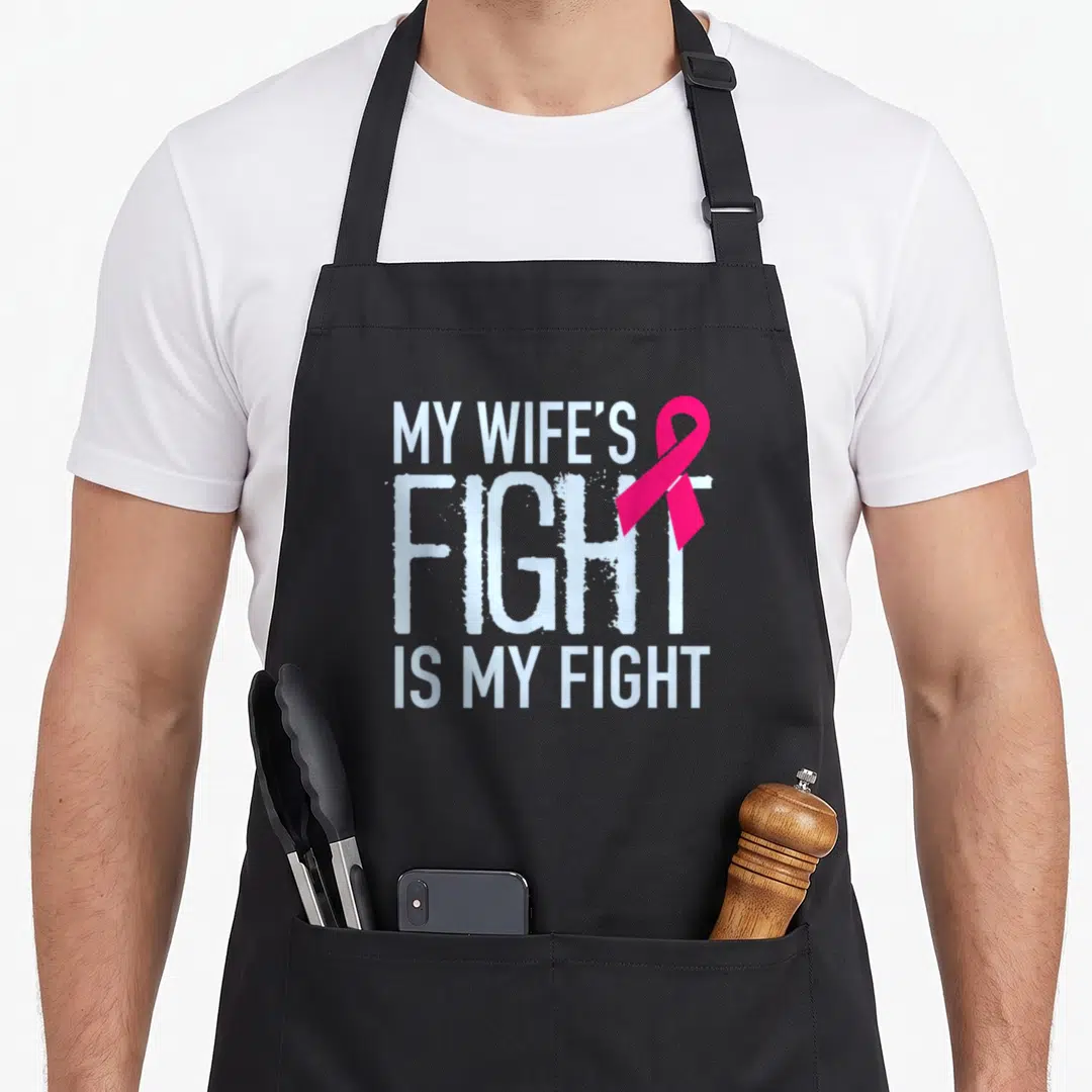 Fight wife apron - White text reading MY WIFE'S FIGHT IS MY FIGHT with overlapping pink ribbon — shown on a black cotton canvas apron against a white background