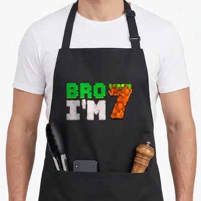 Pixel birthday apron - Pixelated text BRO I'M 7 in green white and orange colors — shown on a black cotton canvas apron against a white background