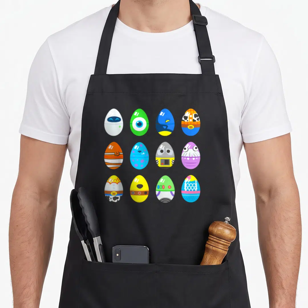 Character easter egg apron - Twelve colorful cartoon eggs in a grid pattern with character details like cow print — shown on a black cotton canvas apron against a white background