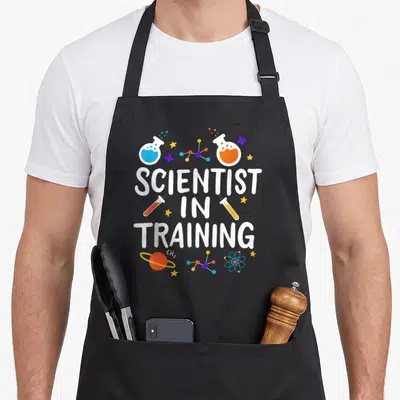 School science apron - Stacked Scientist in Training text with cartoon flasks, molecules, stars, and planet in white — shown on a black cotton canvas apron against a white background