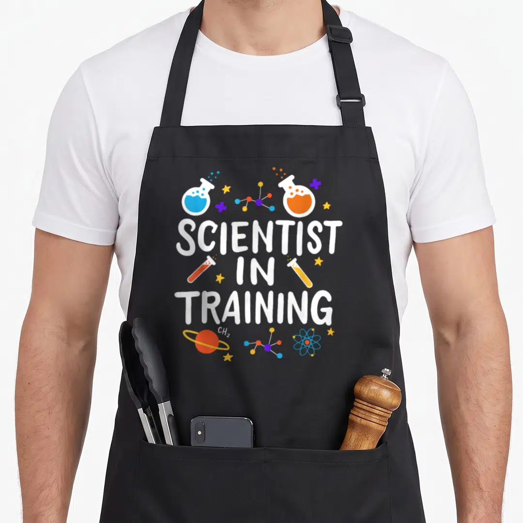 School science apron - Stacked Scientist in Training text with cartoon flasks, molecules, stars, and planet in white — shown on a black cotton canvas apron against a white background