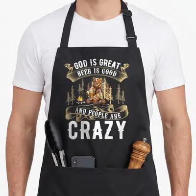 Retro god beer apron - Brown bear holding beer mugs by campfire with white ribbon text — shown on a black cotton canvas apron against a white background